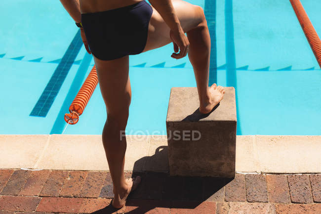 Section basse du nageur mâle debout avec un pied sur le bloc de démarreurs à la piscine extérieure le jour ensoleillé — Photo de stock