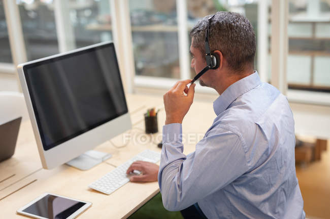 Vista lateral del hombre de negocios que trabaja con computadora y auriculares en el escritorio en la oficina - foto de stock