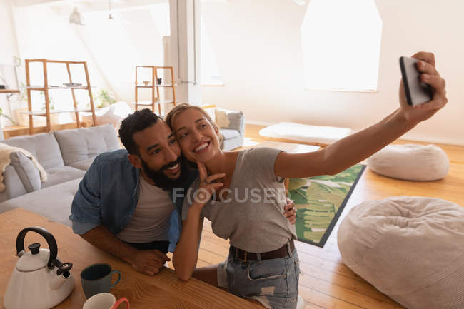 Вид смешанной пары, делающей селфи в гостиной дома — стоковое фото