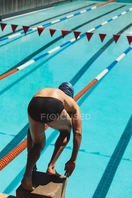 Hochwinkelaufnahme eines männlichen kaukasischen Schwimmers, der auf dem Startblock in Startposition am Schwimmbad in der Sonne steht — Stockfoto