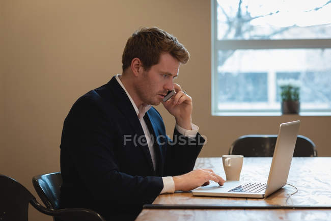 Vista lateral do homem de negócios que trabalha sobre o portátil ao falar no telefone na conferência do escritório — Fotografia de Stock