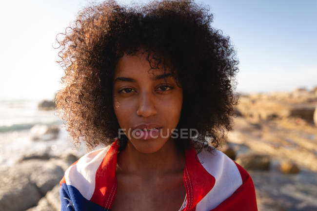 Vista frontal da mulher afro-americana de pé e envolto bandeira americana perto do lado do mar — Fotografia de Stock