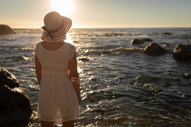 Вид ззаду на активну старшу жінку, дивлячись на морський горизонт, стоячи на березі моря ввечері з великим заходом сонця, що сяє — стокове фото