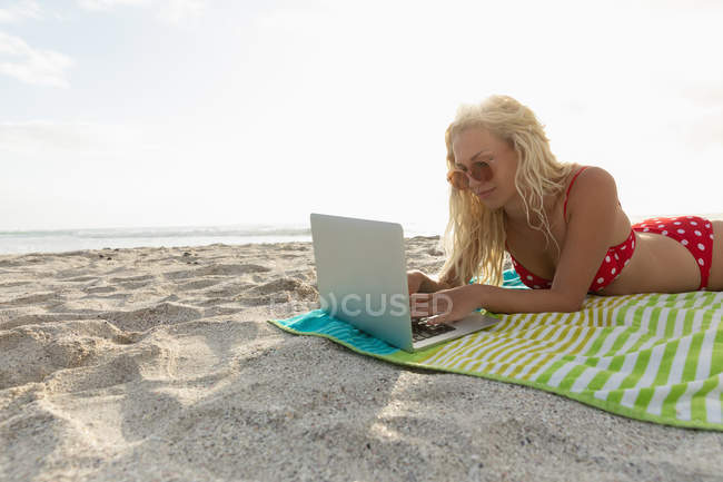 Seitenansicht einer entspannten schönen Frau mit Laptop, während sie an einem sonnigen Tag am Strand liegt. sie arbeitet — Stockfoto