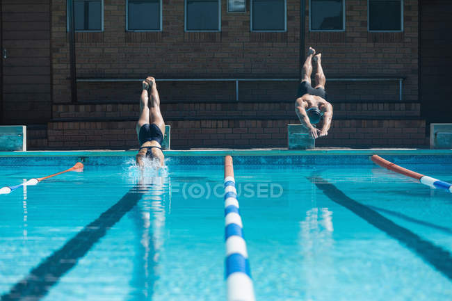 Vista frontal de nadadores caucásicos masculinos y femeninos saltando al agua al mismo tiempo en la piscina bajo el sol - foto de stock