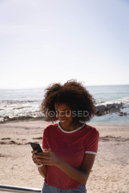 Front view of African-american beautiful woman smiling while using mobile phone at beach — Stock Photo