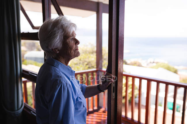 Seitenansicht einer nachdenklichen aktiven Seniorin, die im Schlafzimmer steht und nach draußen schaut — Stockfoto