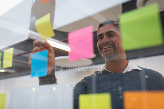 Vue de face d'un homme d'affaires heureux souriant et regardant les notes collantes fixées sur le mur dans le bureau — Photo de stock