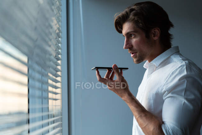 Вид сбоку на молодого руководителя мужского пола разговаривающего по мобильному телефону у окна в современном офисе — стоковое фото
