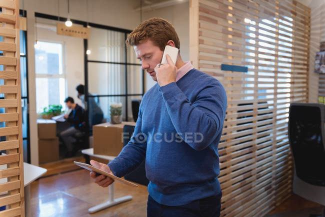 Вид сбоку на бизнесмена, использующего цифровой планшет во время разговора по мобильному телефону в офисе — стоковое фото