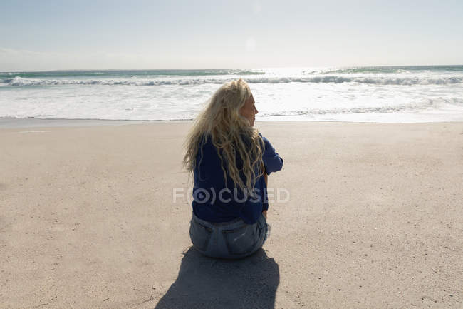 Visão traseira da bela mulher loira sentada na praia em um dia ensolarado. Ela está olhando a paisagem — Fotografia de Stock