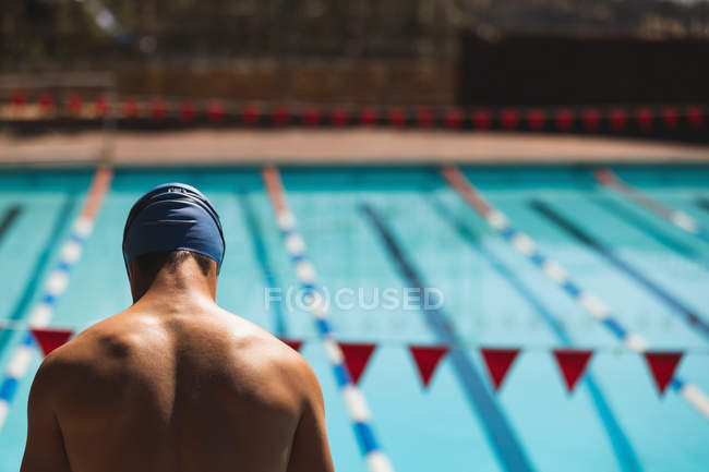 Vue arrière du jeune nageur masculin caucasien regardant vers le bas tout en se tenant à la piscine au soleil — Photo de stock
