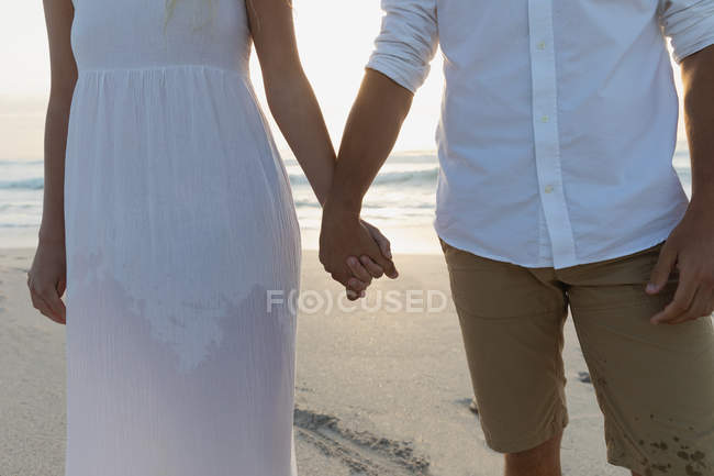 Der mittlere Teil des jungen Liebespaares hält Händchen, während es an einem sonnigen Tag am Strand steht. Sie genießen ihren Urlaub — Stockfoto