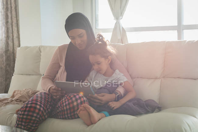 Vue de face de mère mixte portant hijab et fille utilisant une tablette numérique dans le salon à la maison — Photo de stock