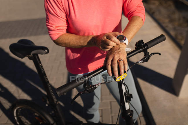 Средняя часть активной пожилой женщины, использующей свои умные часы, держа свой велосипед на прогулке под солнцем — стоковое фото