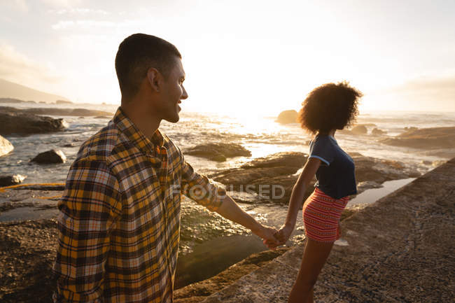 Vista lateral de una pareja afroamericana cogida de la mano y parada en la roca cerca del lado del mar - foto de stock