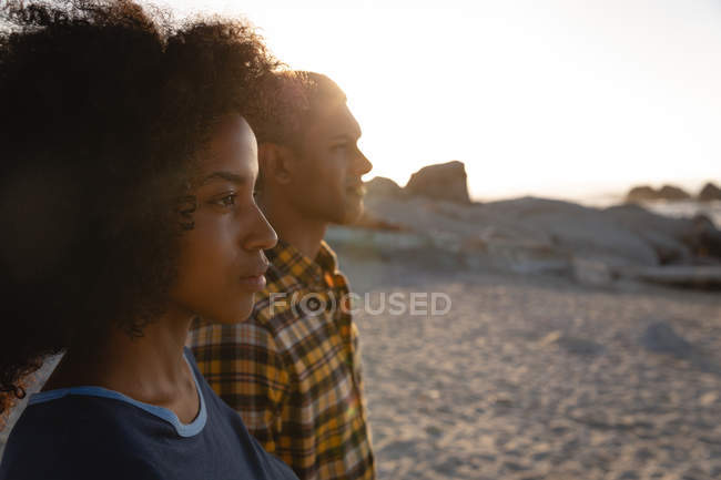 Вид сбоку на афро-американскую пару, стоящую на пляже. Они так же смотрят на закат — стоковое фото