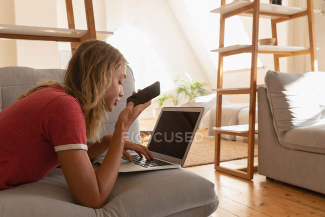 Vista lateral de la mujer que usa el ordenador portátil mientras habla en el teléfono móvil en la sala de estar en casa - foto de stock