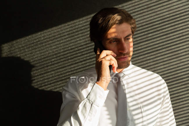 Вид спереду красивий молодий чоловік-виконавця розмовляє на мобільному телефоні, стоячи біля вікна в сучасному офісі — стокове фото