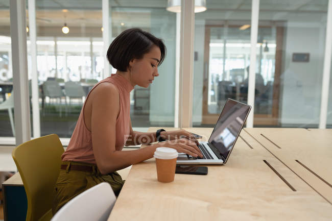 Vista lateral de una mujer de negocios asiática usando un portátil en el escritorio en la oficina - foto de stock