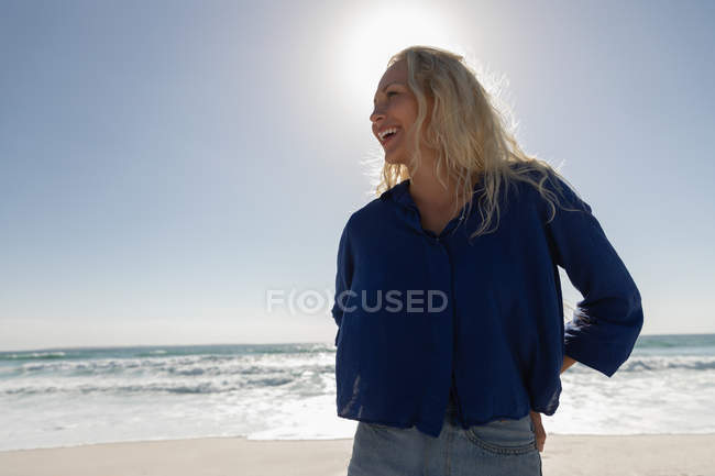 Vista frontal da bela mulher loira sorrindo enquanto está de pé na praia em um dia ensolarado. Ela está sorrindo. — Fotografia de Stock