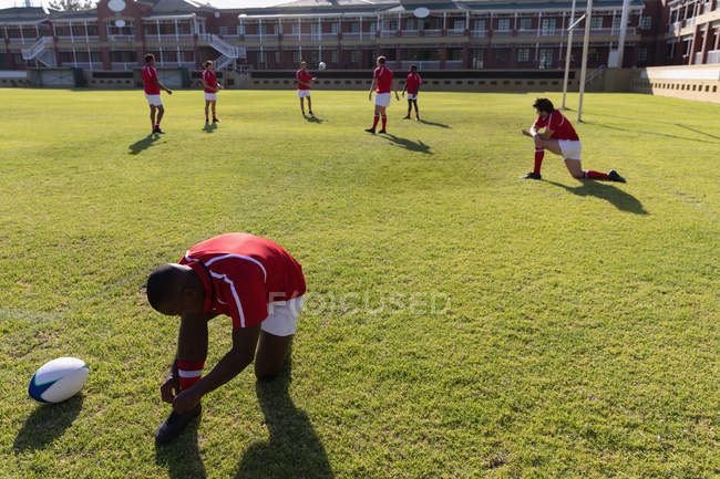 Vista frontale del giocatore di rugby afroamericano che lega i lacci delle scarpe sul campo da rugby con i membri della squadra sullo sfondo nella giornata di sole — Foto stock