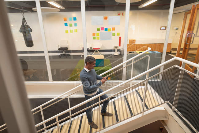Высокий угол обзора бизнесмена с помощью мобильного телефона во время восхождения по лестнице в офисе — стоковое фото