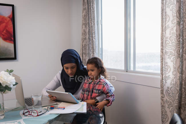Vista frontal da mãe parda vestindo hijab e filha usando tablet digital em casa. Eles estão sentados em torno de uma mesa na sala de estar — Fotografia de Stock