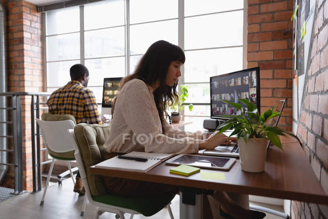 Seitenansicht einer kaukasischen Geschäftsfrau, die über Laptop arbeitet, während ein afrikanisch-amerikanischer Mann hinter ihr im Büro arbeitet — Stockfoto