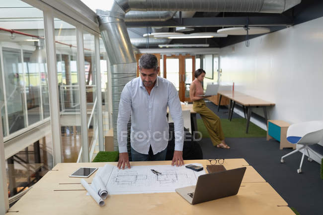 Vista frontale di Businessman in piedi in ufficio e lavorare su un progetto alla scrivania mentre una donna d'affari lavora su un computer portatile in background — Foto stock