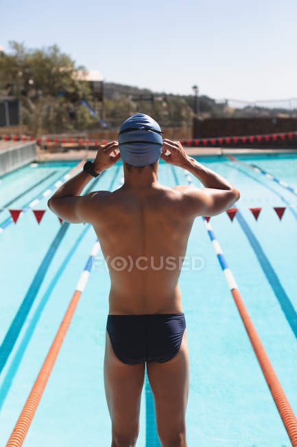 Vista trasera de un joven nadador caucásico ajustando las gafas de natación mientras está parado en la piscina al aire libre en un día soleado - foto de stock