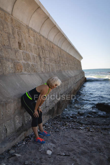 Side view of an active senior woman taking a break of her exercise against a wall on the beach — Stock Photo