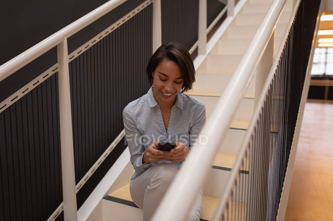 Vista frontal de uma empresária asiática usando seu telefone celular enquanto sentada nas escadas do escritório — Fotografia de Stock