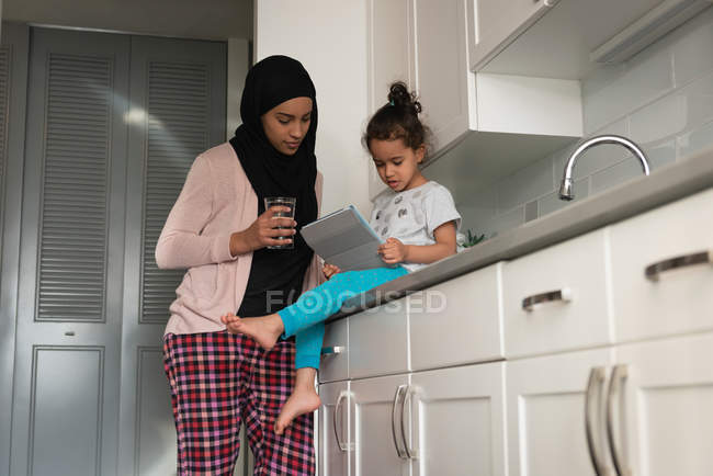 Низкий угол обзора матери смешанной расы, надевающей хиджаб и дочери вместе, используя цифровой стол в кухонной комнате дома — стоковое фото