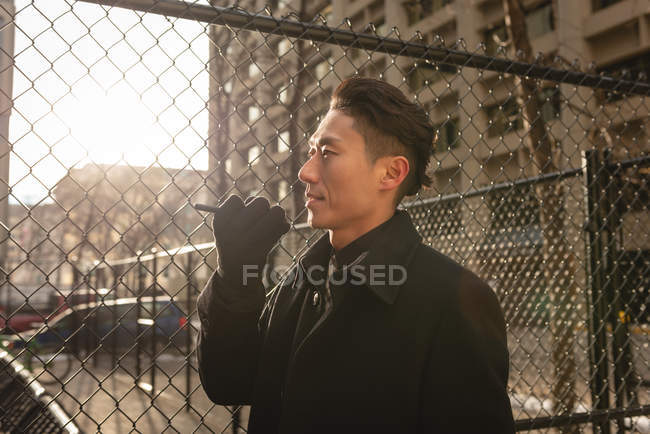 Side view of young Asian businessman talking on mobile phone while walking past chain link fence on street in the city — Stock Photo