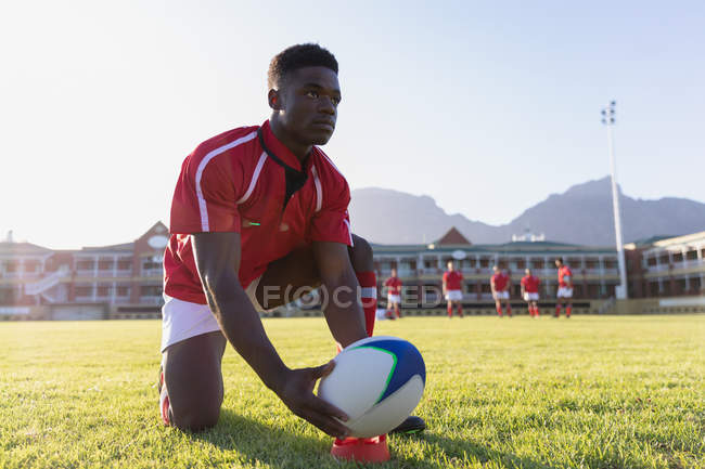 Vista frontal de un joven jugador de rugby afroamericano que coloca la pelota de rugby al patear tee en el estadio - foto de stock