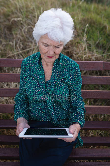 Vista frontal de una mujer mayor activa usando una tableta digital mientras está sentada en un banco de madera en la playa - foto de stock