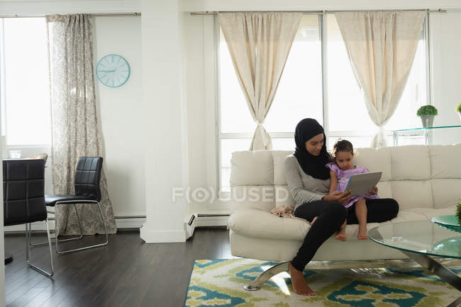 Vista frontal de madre de raza mixta usando hijab e hija usando tableta digital en la sala de estar en casa - foto de stock