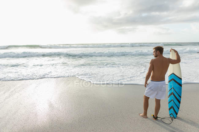 Vista posteriore del surfista biondo maschio con una tavola da surf in piedi sulla spiaggia in una giornata di sole. Sta guardando le onde — Foto stock