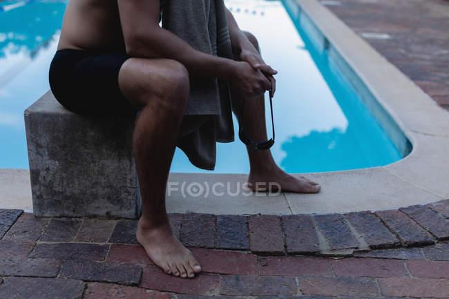 Sección baja de un nadador masculino sentado en el bloque de salida cerca de la piscina - foto de stock