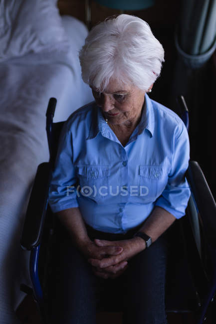 Blick aus der Vogelperspektive auf eine gehbehinderte Seniorin mit zusammengeklappten Händen, die im heimischen Schlafzimmer auf einem Rollstuhl sitzt — Stockfoto