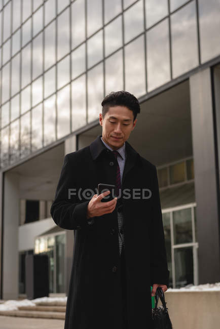 Front view of young well dressed Asian businessman looking at mobile phone while walking on street in the city — Stock Photo