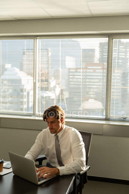 Вид спереди красивого молодого мужчины-руководителя, сидящего за столом и работающего на ноутбуке в современном офисе — стоковое фото