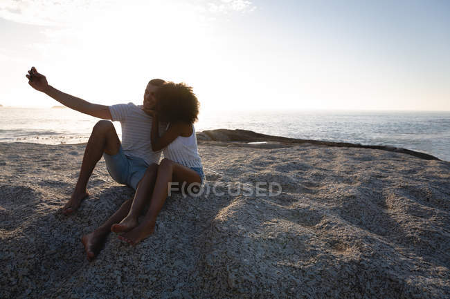 Vorderansicht eines afrikanisch-amerikanischen Paares, das ein Kuss-Selfie macht, während es in der Nähe des Meeres sitzt — Stockfoto