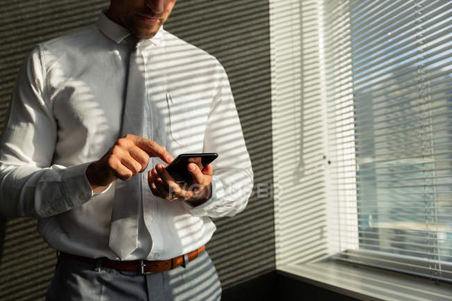 Средняя секция красивого молодого мужчины, работающего на мобильном телефоне, стоя у окна в современном офисе — стоковое фото