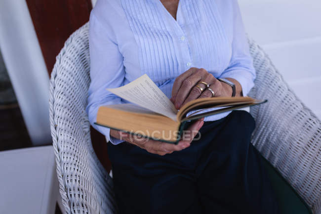 Средняя часть активной пожилой женщины, читающей книгу, сидя дома на кресле на балконе — стоковое фото