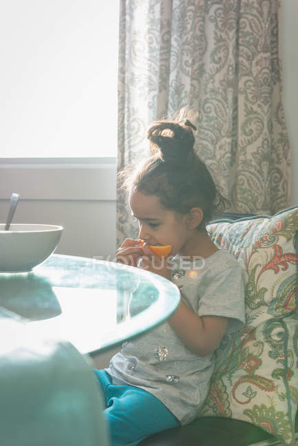 Девочка смешанной расы ест апельсиновый фрукт, сидя на стуле дома — стоковое фото
