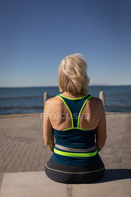 Rückansicht einer aktiven Seniorin, die es sich auf einer Bank an der Promenade gemütlich macht und in der Sonne aufs Meer blickt — Stockfoto