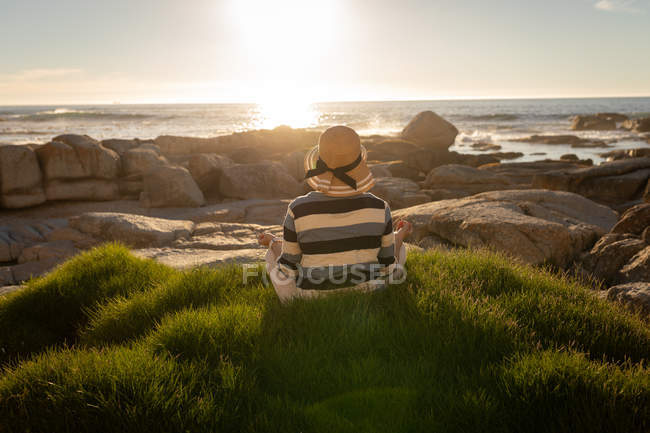 Rückansicht einer aktiven Seniorin, die vor Sonnenuntergang Yoga macht, während sie am Strand auf Gras sitzt — Stockfoto