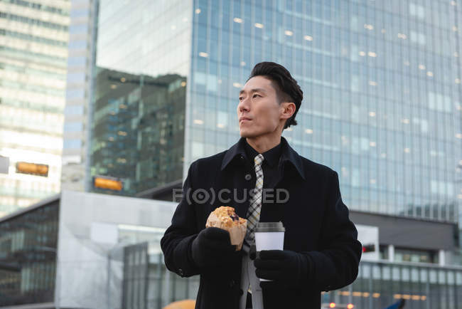Front view of young Asian businessman thinking while standing on street in the city. Holding coffee and bakery — Stock Photo